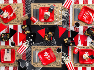 table decorations with square red plates and red cups with skull and bone white picture, brown and black illustration treasure map place mats. Black and white striped paper napkins in black and white with red triangle bunting. Pirate themed birthday party for girls and boys party or birthday parties from la di dah