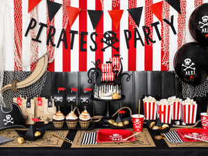 table decorations with square red plates and red cups with skull and bone white picture, brown and black illustration treasure map place mats. Black and white striped paper napkins in black and white with red and black triangle bunting. Black Pirate party hanging banner, pirate party bags in brown paper, sails in red and white cake toppers with black octopus legs. Pirate themed birthday party for girls and boys party or birthday parties from la di dah